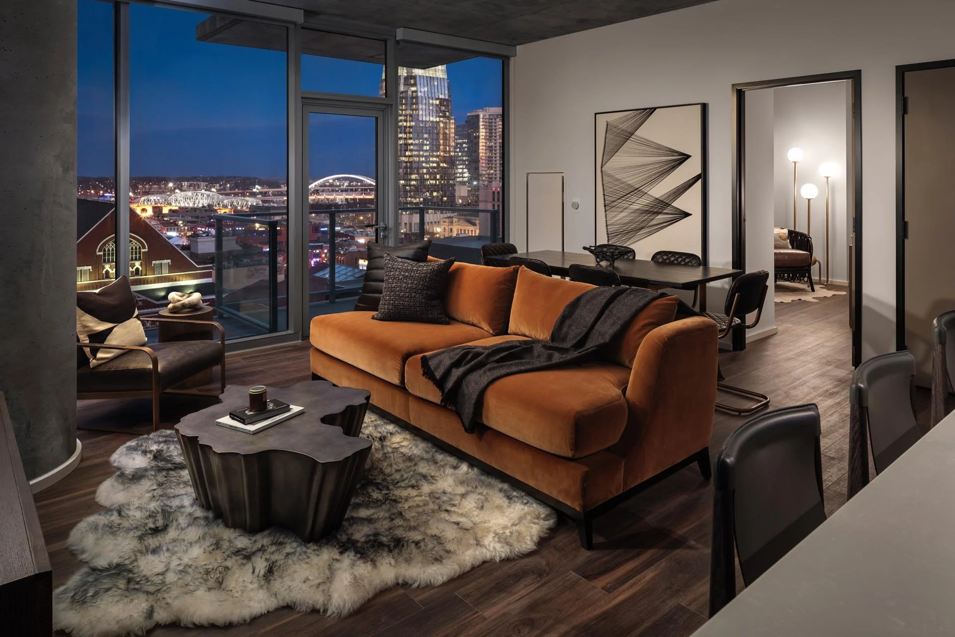 Modern living room in an apartment with large windows showing a city skyline at night, including a bridge and tall buildings, furnished with a burnt orange sofa, black armchair, coffee table, and a sheepskin rug.
