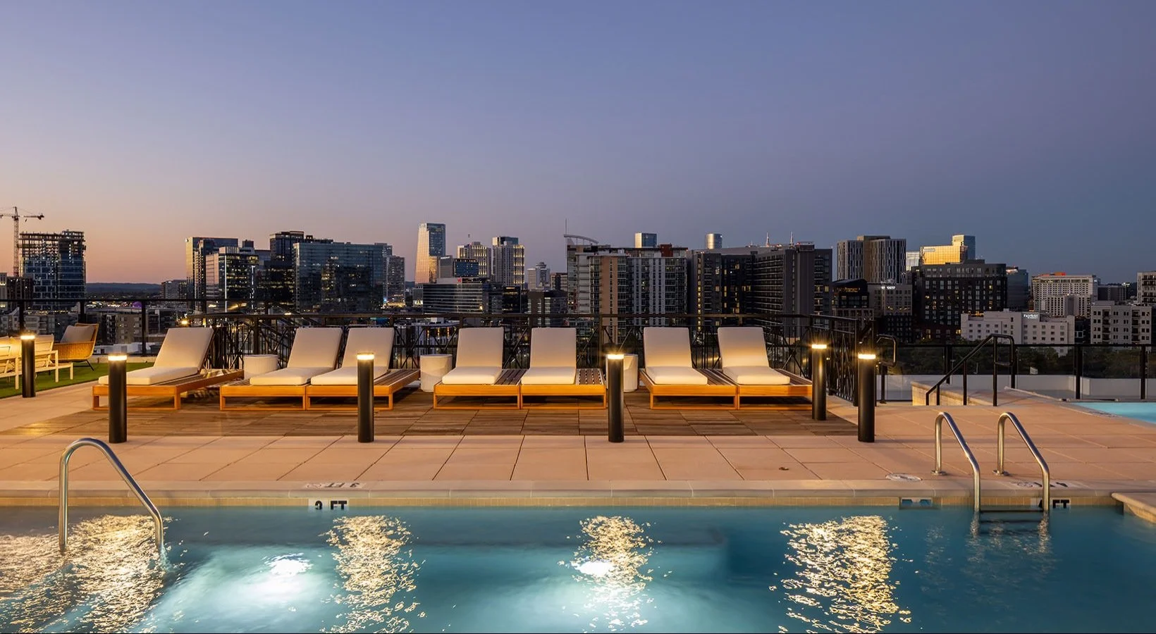 Urban rooftop pool area with lounge chairs, city skyline in the background at dusk.