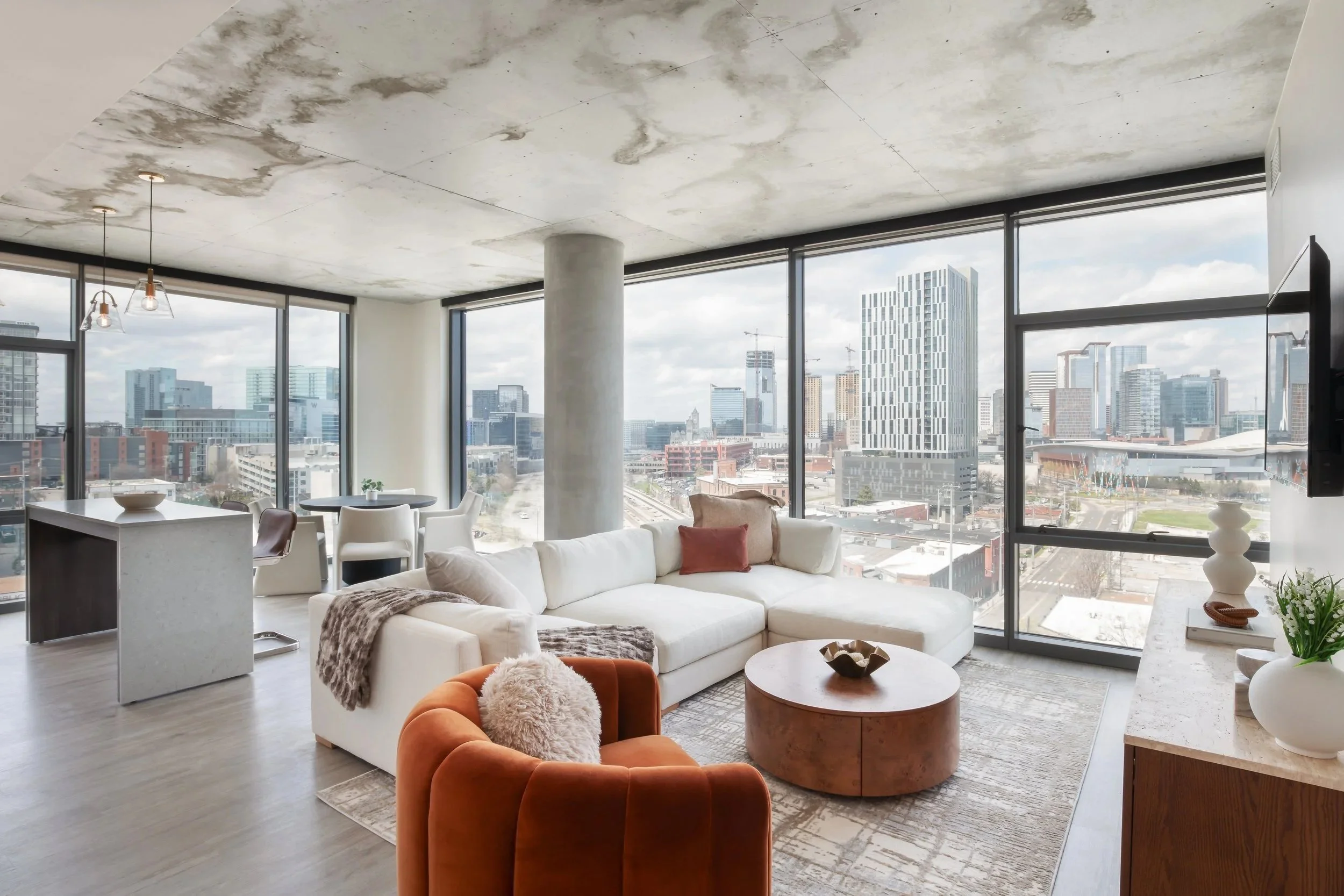 Modern living room with large windows overlooking a cityscape, white sectional sofa with decorative pillows, orange accent chair, round wooden coffee table, a dining area with white chairs and a small table, and contemporary decor including vases and a sculpture.