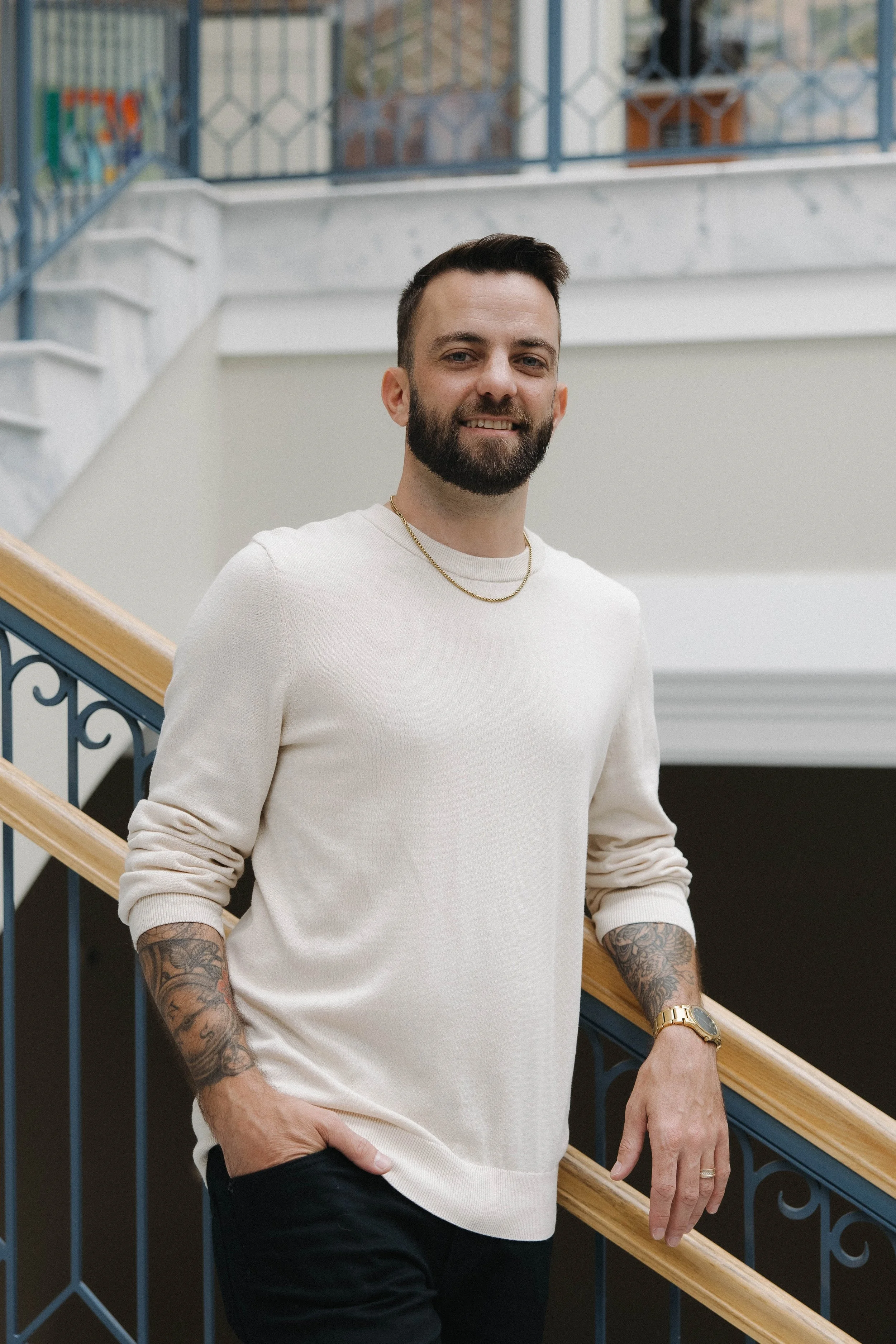 A man with a beard and short dark hair, wearing a cream-colored sweater, black pants, a gold watch, and a gold chain, stands on a staircase inside a building, smiling at the camera.
