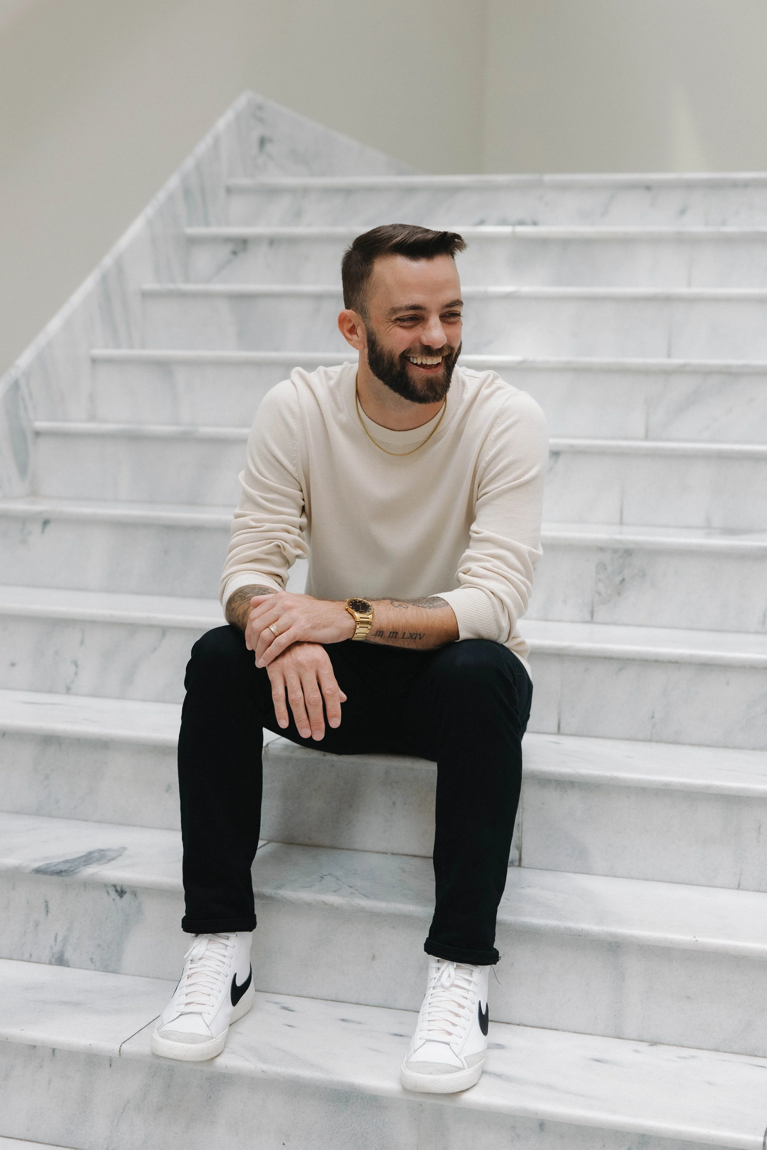 A man with a beard, wearing a white sweatshirt, black pants, and white sneakers with a black swoosh, sitting on marble stairs, smiling and looking to his right.