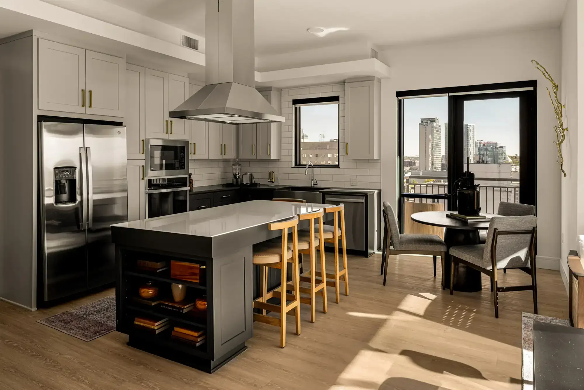 Modern kitchen with white cabinets, black lower cabinets, stainless steel refrigerator, microwave, oven, and a large white island with wooden stools. There is a small dining area with a round table, four chairs, and a view of city buildings through large windows and a sliding glass door.