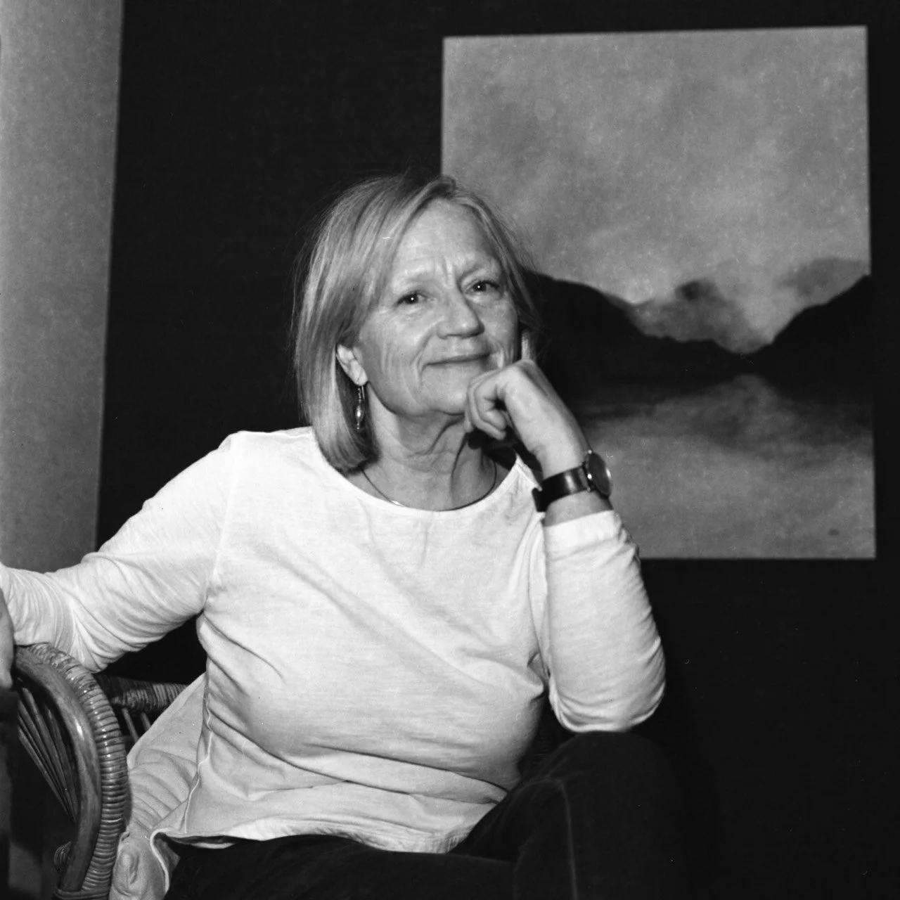 A smiling older woman with short hair, wearing a white top, sitting indoors in front of a landscape photograph.
