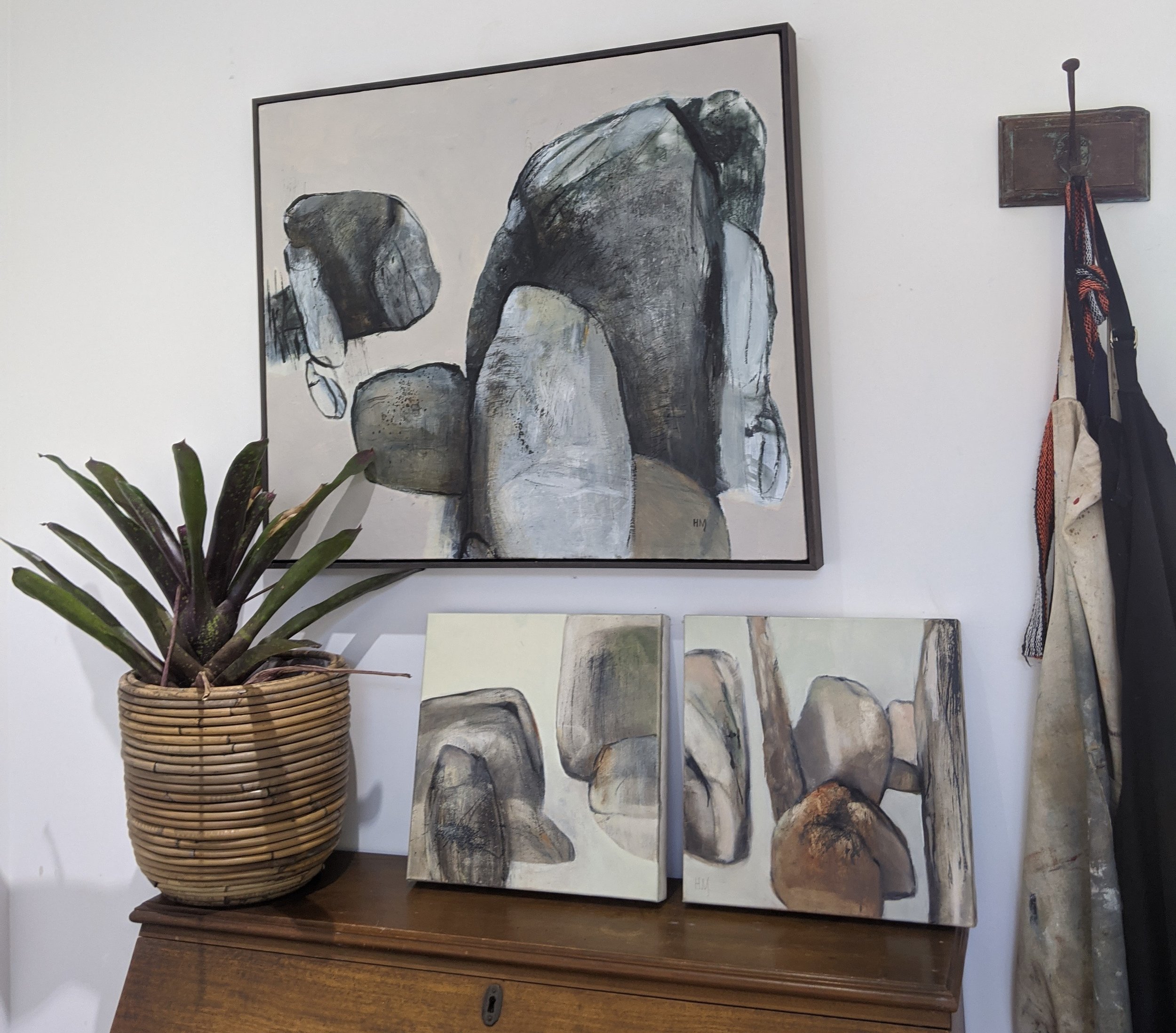Wall art depicting abstract painting of rocks, displayed above a wooden cabinet with a potted plant, and two small canvas paintings of rocks resting on the cabinet. Clothes hanging on hooks on the wall to the right.