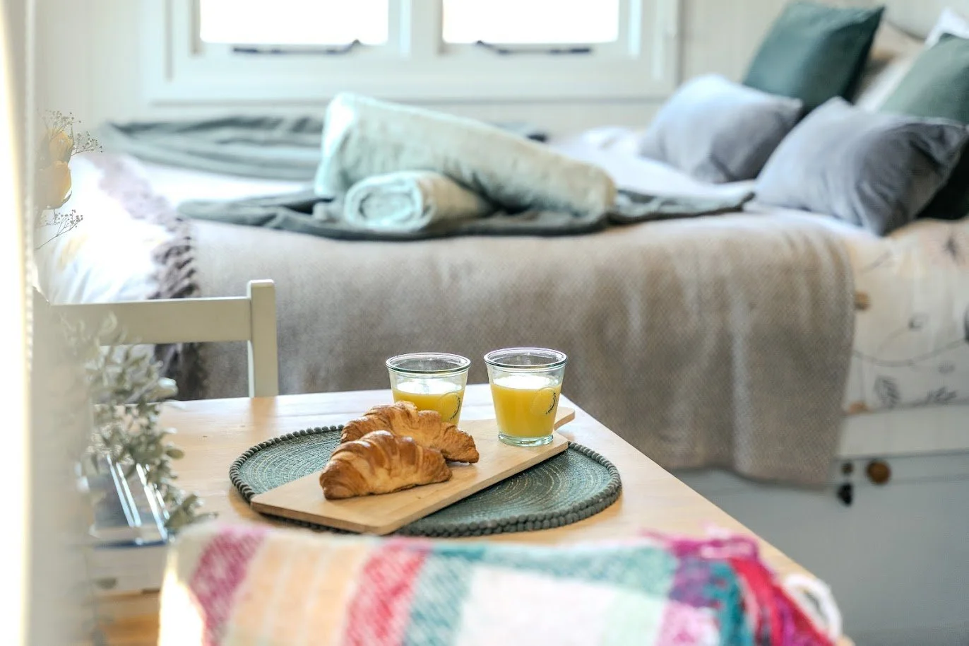 A breakfast table with two croissants and two glasses of orange juice, set with a green placemat and a colorful cloth, in a cozy room with a bed and pillows in the background.