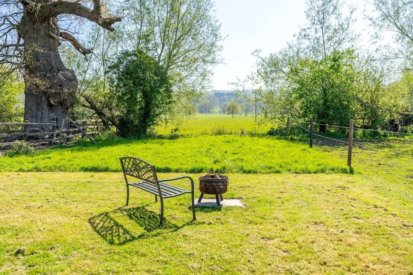 A small metal park bench and a fire pit on a grassy yard with trees and a fence, overlooking a field and distant trees under a clear sky.