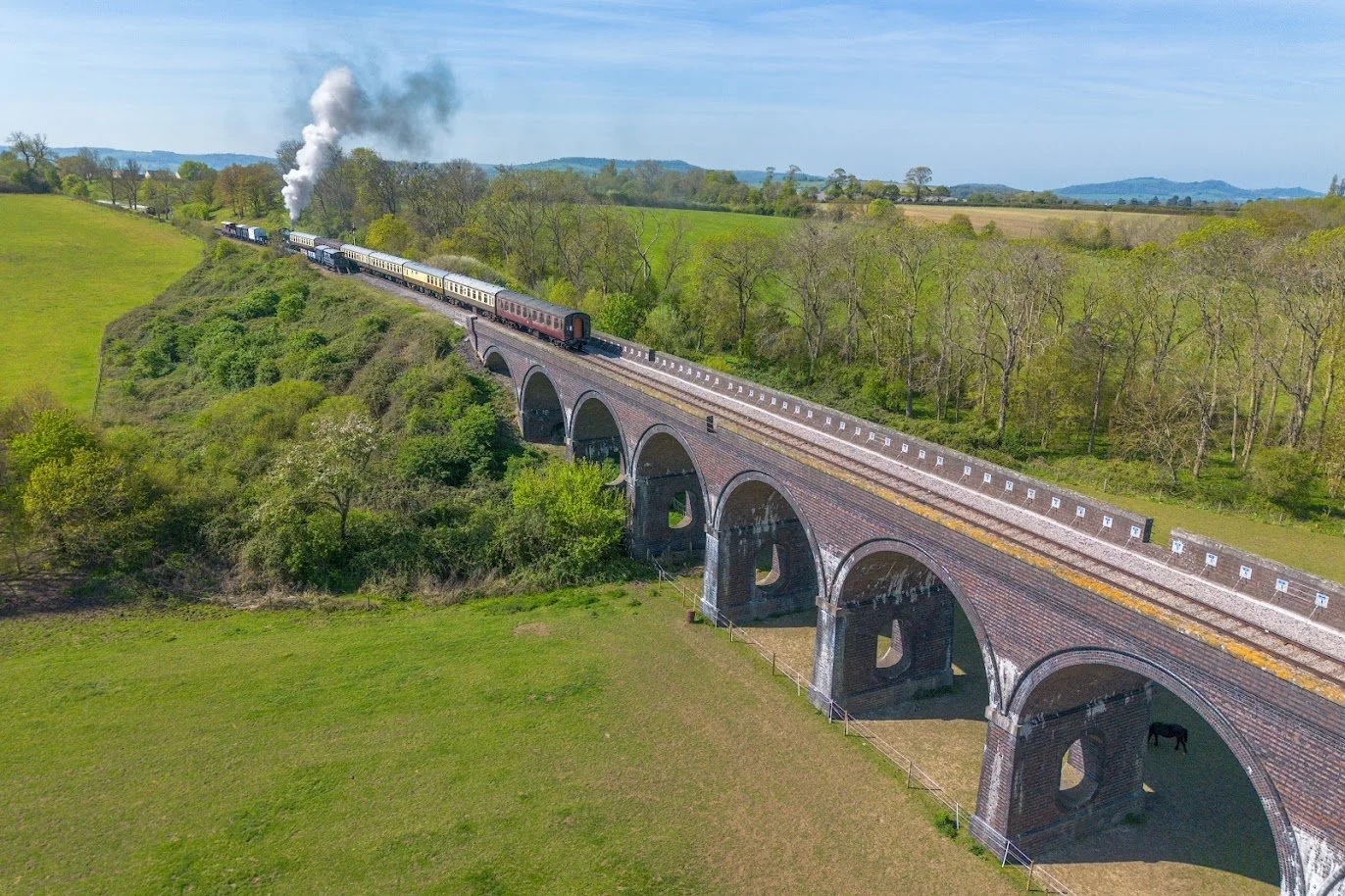 A vintage steam train traveling over a historic arched stone bridge through lush, green countryside during daytime.