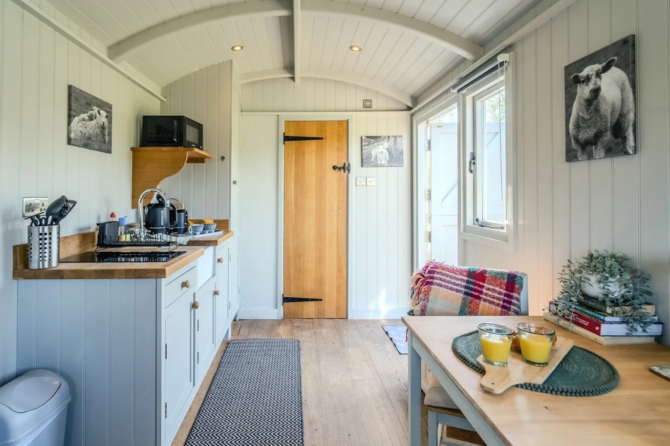 Cozy small kitchen with white paneled walls, wooden countertops, a microwave, and kitchen utensils. There is a window and door letting in natural light, with framed animal photographs on the walls. A dining table with two glasses of orange juice, boo