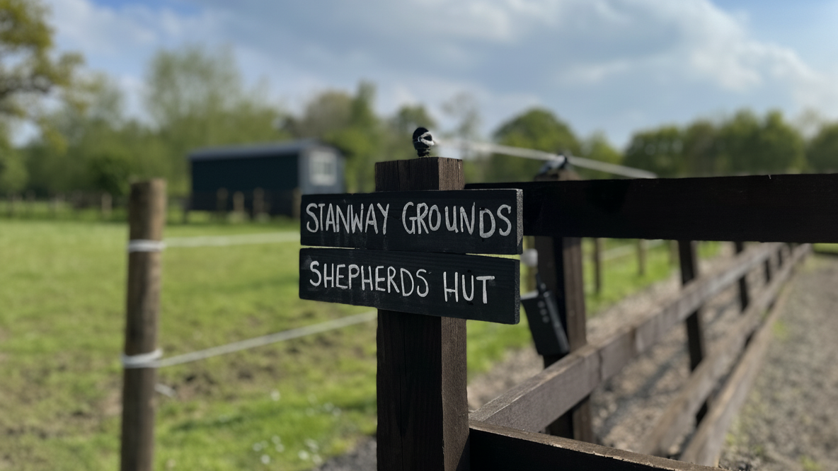 A wooden sign pointing to Stanway Grounds and Shepherds Hut, with a rural farm in the background and horses grazing in the back ground.