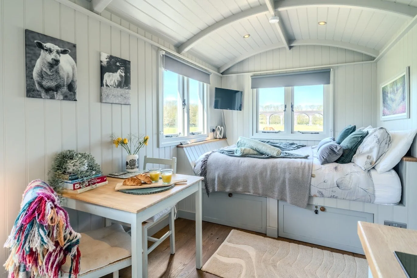 Sunlit cozy bedroom with a built-in bed under windows, decorated with pillows and blankets, a small table with breakfast, and wall art, in a bright, airy space.