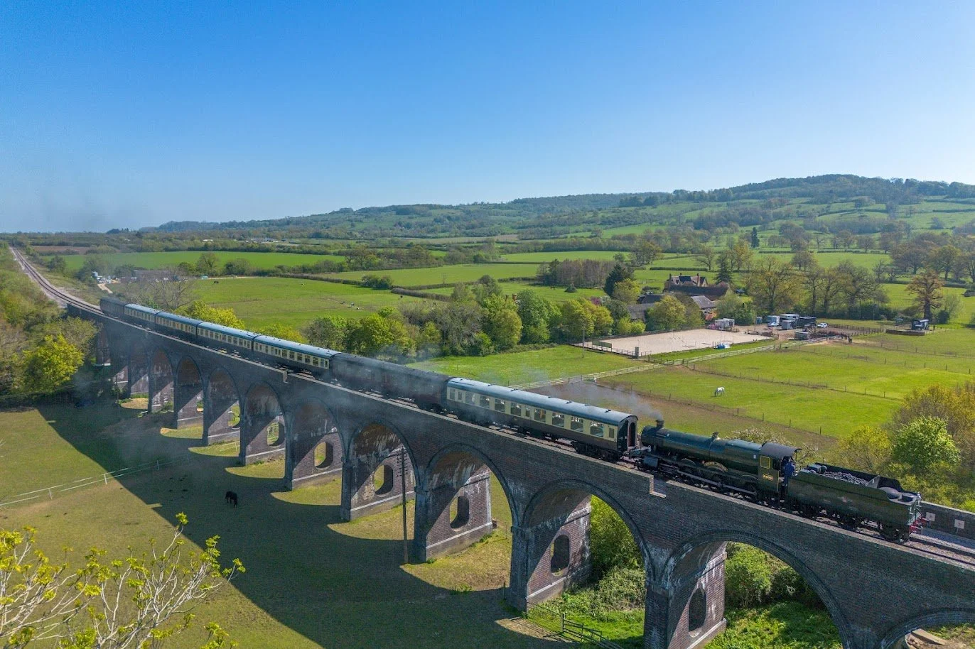 A steam train with passenger cars traveling over a stone arch bridge in a rural landscape with green fields, trees, and hills under a clear blue sky.