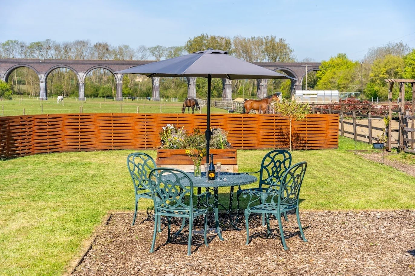 Outdoor patio with a metal table, six chairs, a large umbrella, and a flower arrangement, overlooking a fenced yard with horses grazing and a bridge in the background on a sunny day.