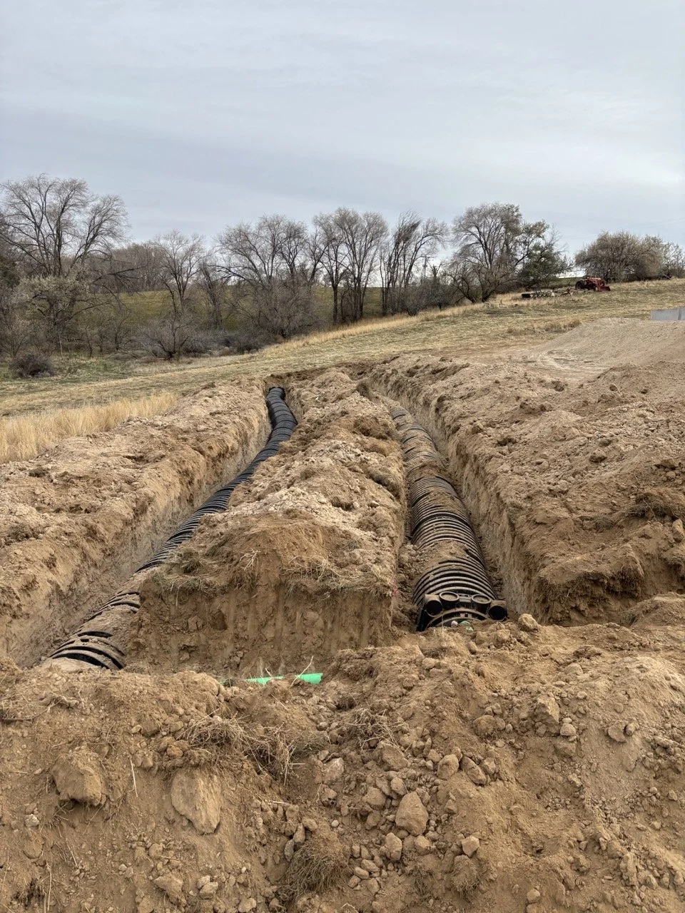 Construction site with two large underground pipes laid in trenches for a septic drain field installation, surrounded by dirt and soil, with trees and a field in the background.
