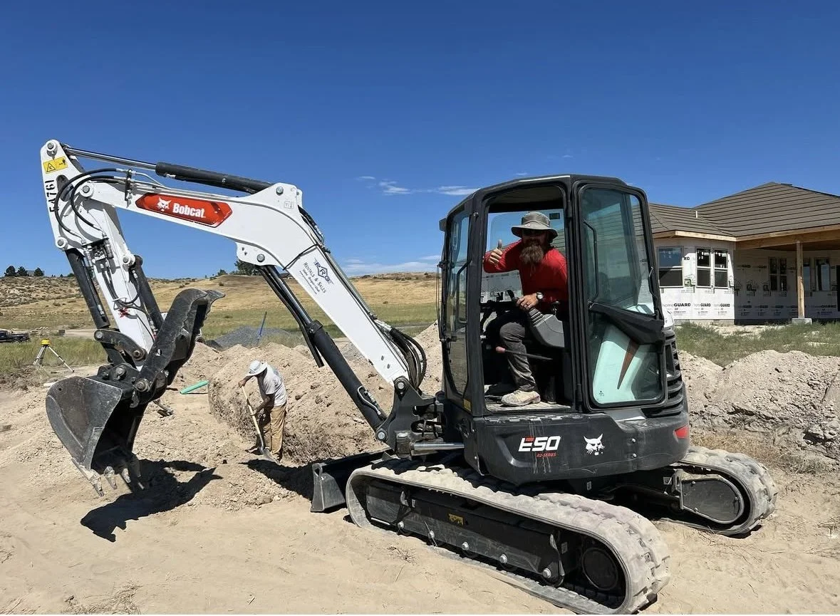 A man with a beard and hat sitting in a small black excavator on a construction site, with another worker in the background digging. A house under construction with a septic drain field repair is visible nearby under a clear blue sky.
