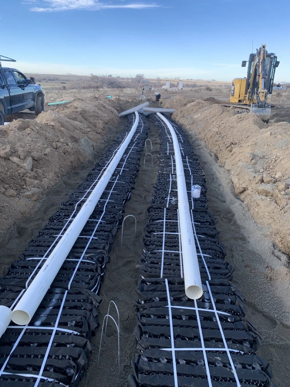 Septic system construction site showing underground piping installation with PVC pipes laid on black fabric and secured with white straps, surrounded by excavated dirt and construction equipment.