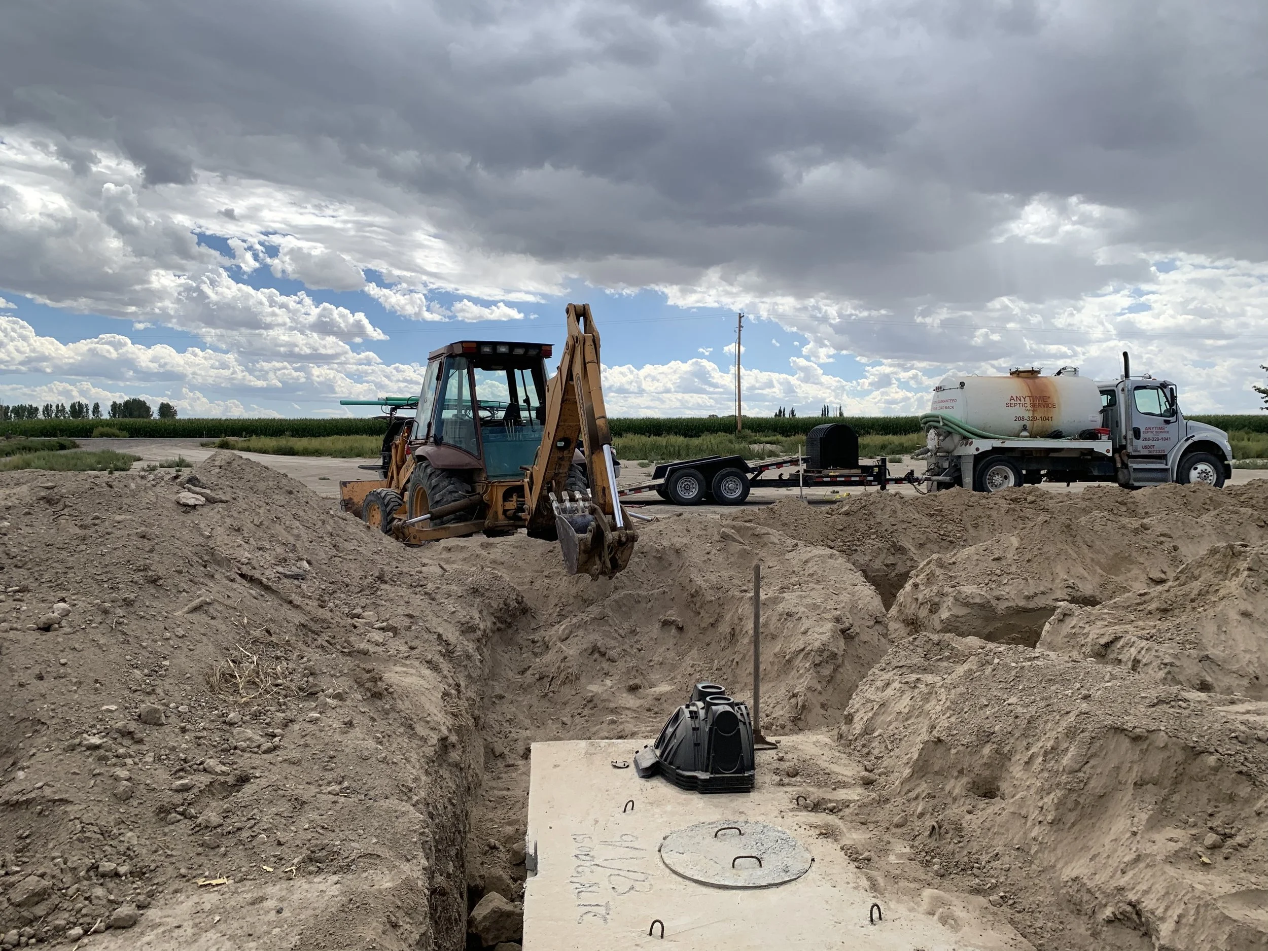 Construction site showing a backhoe, a septic service truck, and a deep trench in the dirt while the are installing a septic tank and drain field under a cloudy sky.
