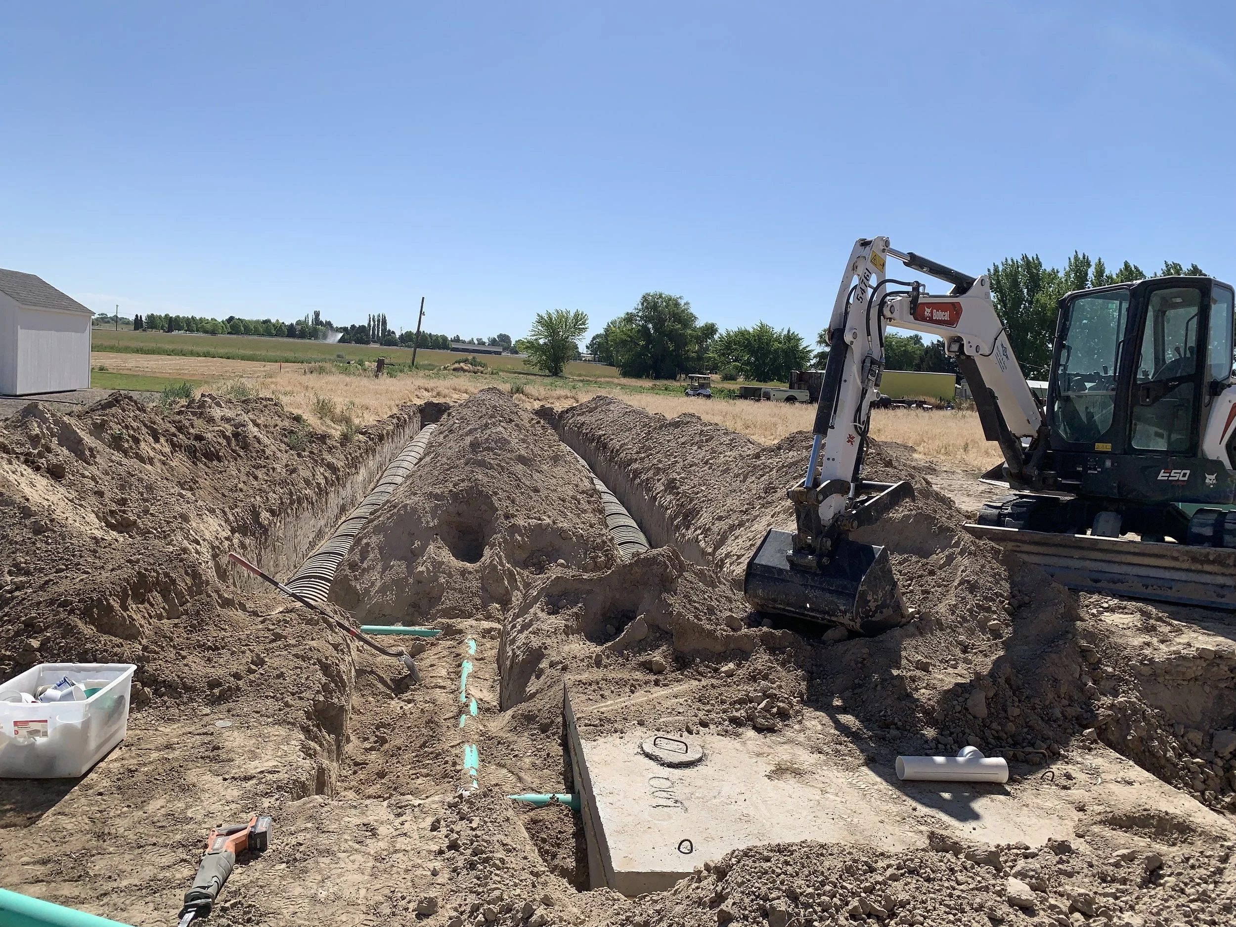 Construction site with an excavator working on underground pipes, septic drain field and septic tank, in a rural area with open fields, a partly cloudy sky, and a small building to the left.