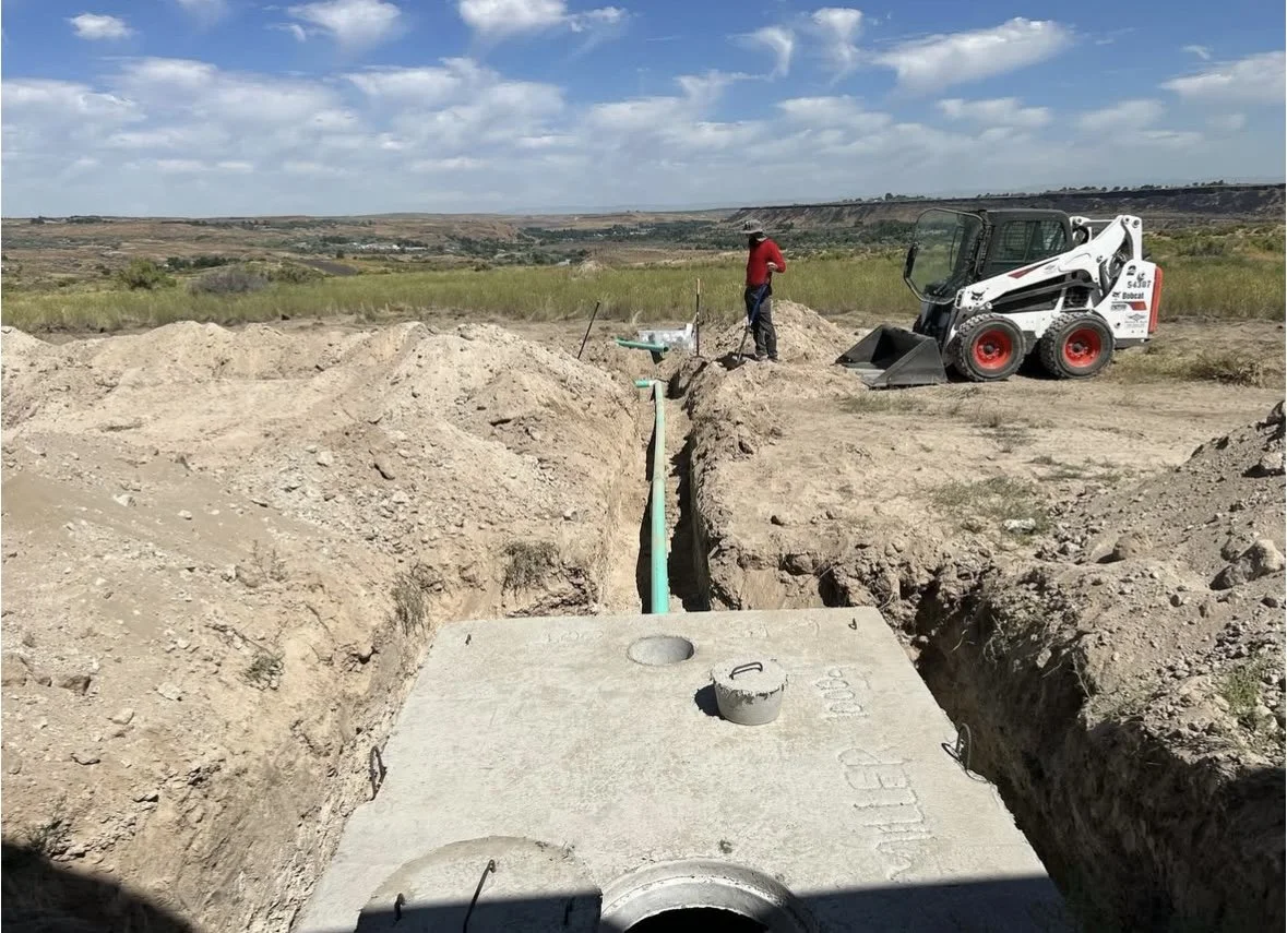 A construction site in a wide open landscape with a worker operating a small Bobcat skid-steer loader near a narrow trench. A green pipe runs through the trench, with a large concrete septic tank and cap visible in the foreground.