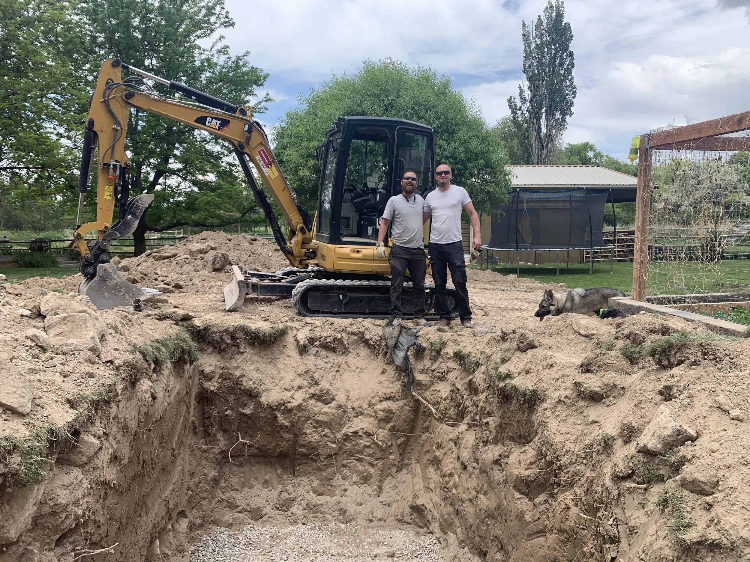 Two men with sunglasses standing on edge of a large hole in the ground, dug for a large septic tank, with a small yellow excavator behind them.