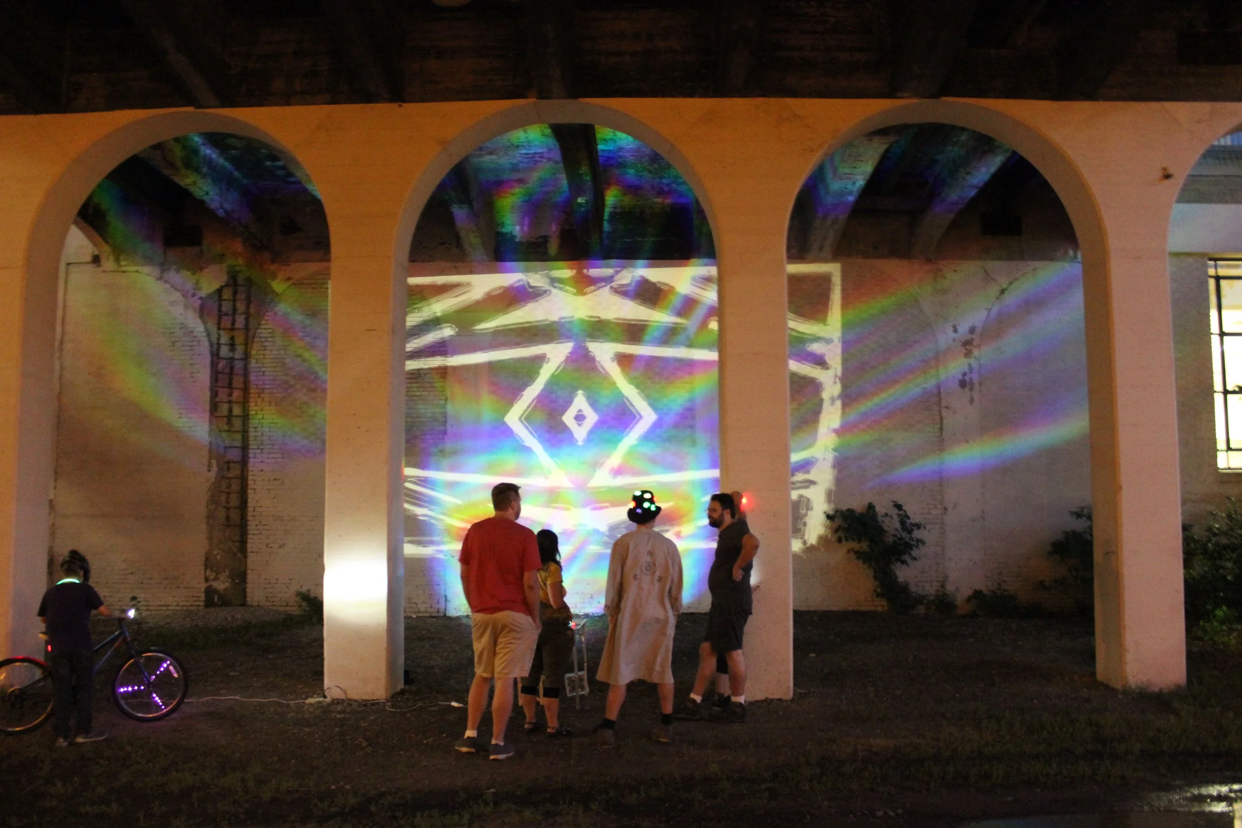 People gathered under a large archway with colorful light projections and laser effects on a wall, some observing the visual display and one person riding a bicycle with lights at night.