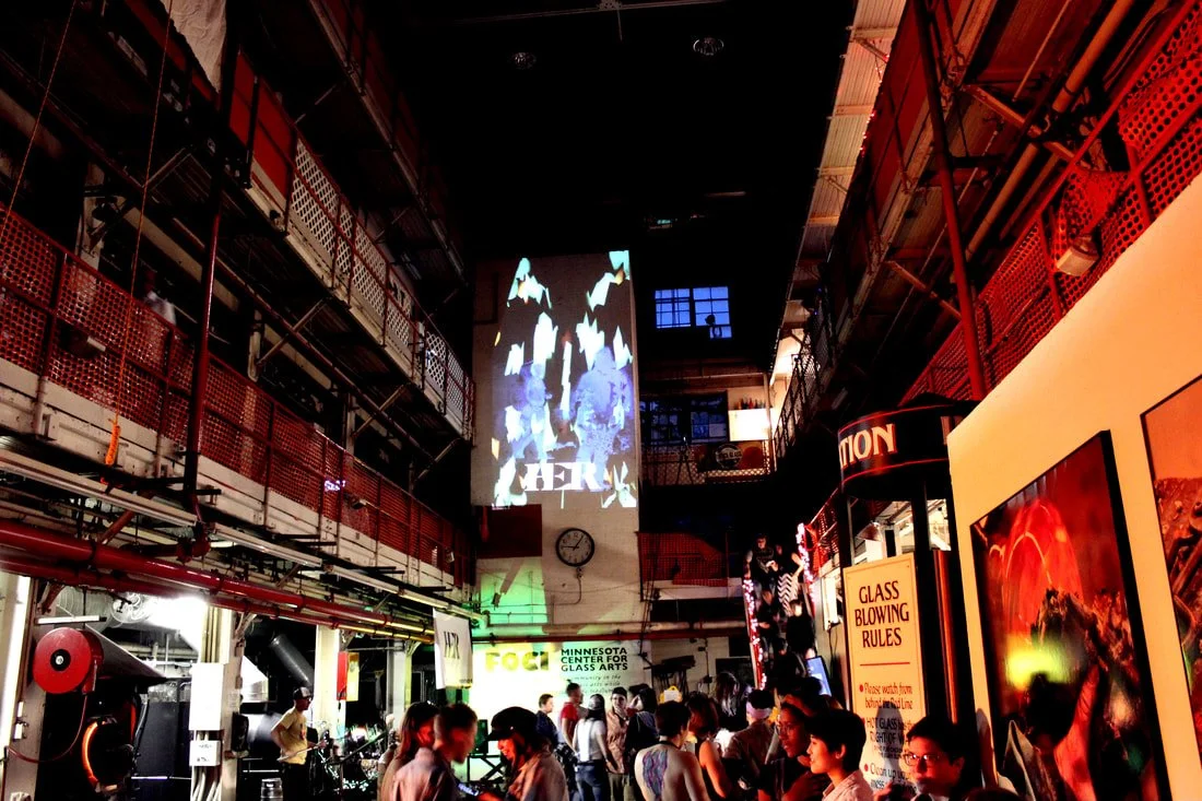Interior of an art gallery or studio with a crowd of people, high ceiling, and industrial decor, including a large projection of artwork on the wall and signs related to glass art.