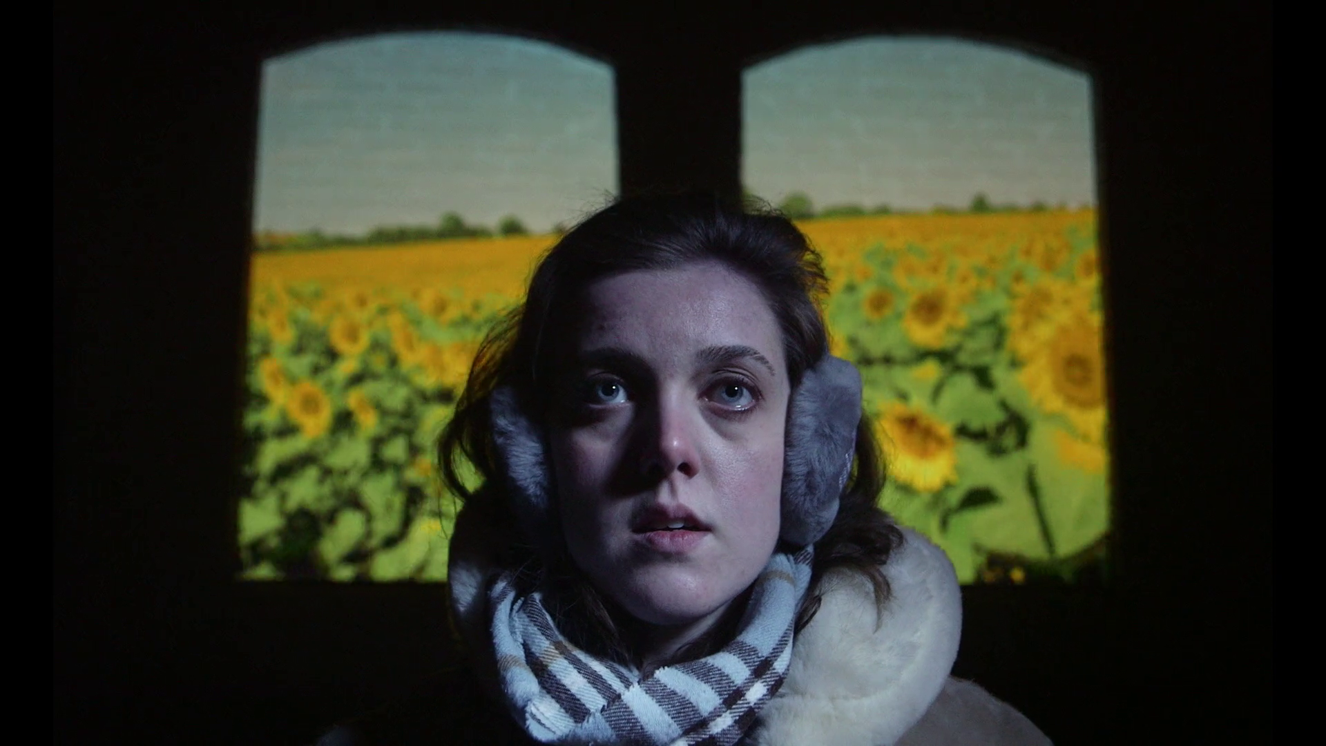 A woman looking upward, wearing earmuffs and a scarf, with a sunflower field in the background.