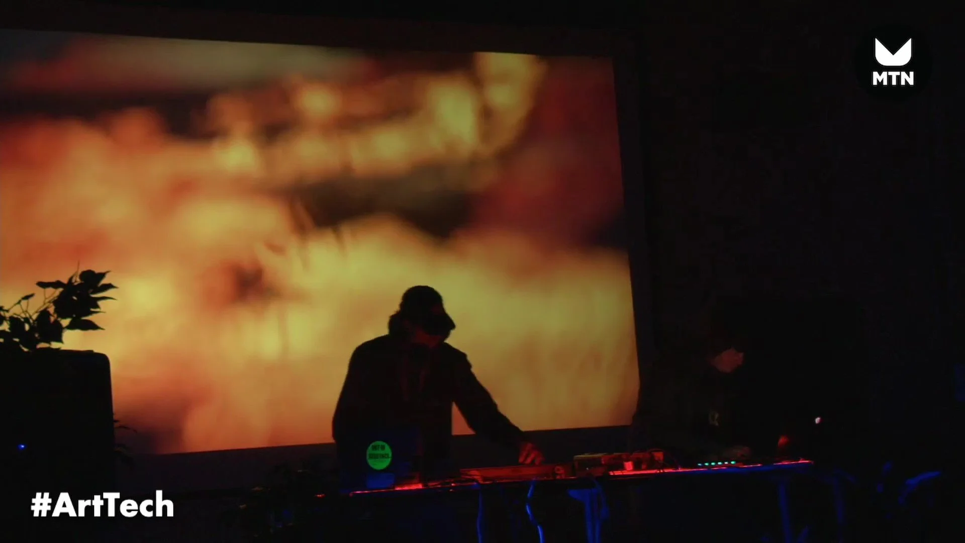 A DJ performing at an event silhouetted against a large, colorful screen with abstract visuals, with some equipment and a plant visible in the foreground.