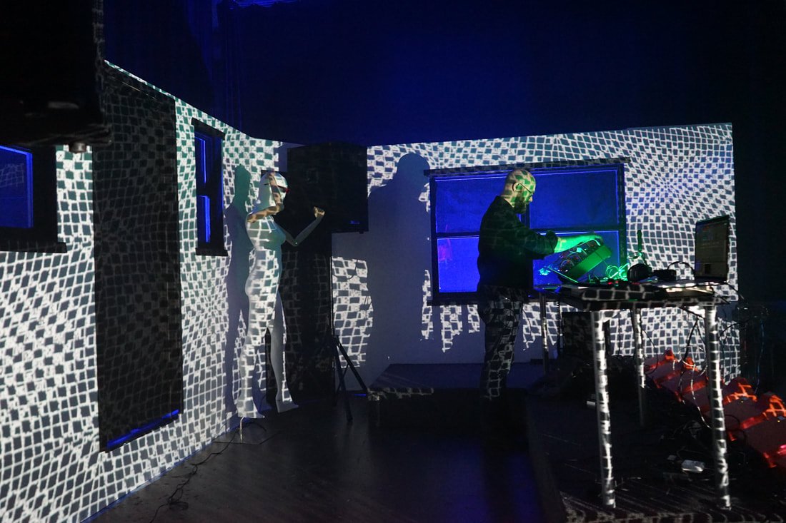 Two people, a DJ and a female singer, performing in a dark room with black and white patterned walls projected with geometric light patterns. The DJ is at a table with electronic equipment, and the female singer stands with a microphone.