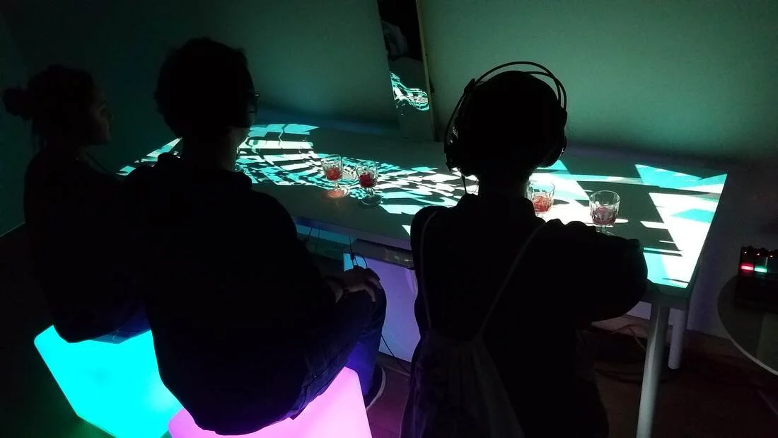 Three people joining an interactive art installation with a digital light display and small glasses of red beverage.