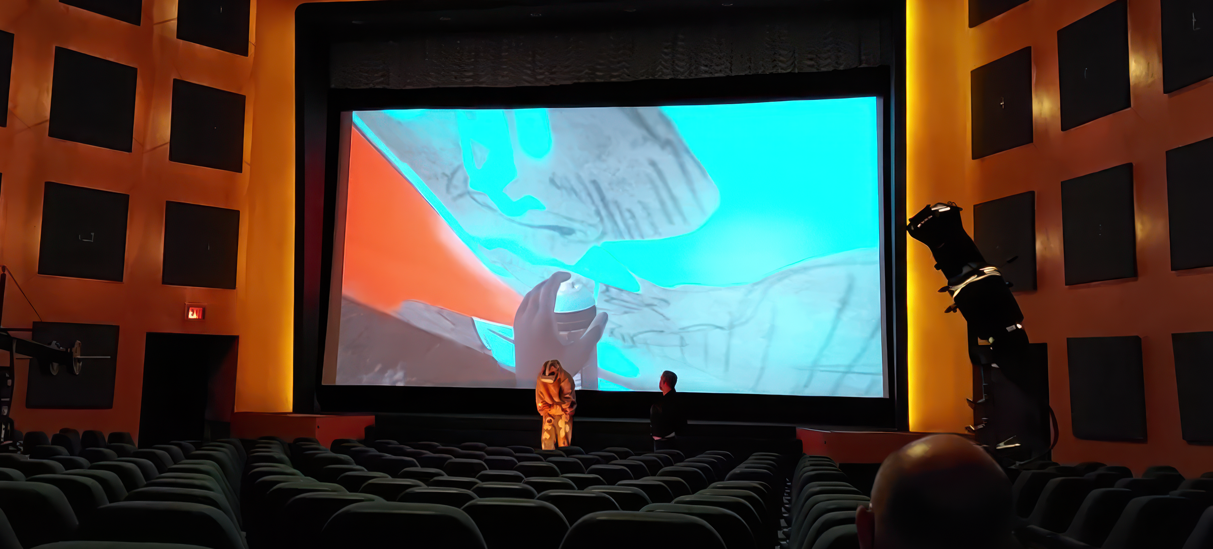A movie theater with a large screen displaying an animated scene of a hand drawing on paper. Two people are standing in front of the screen, looking at the projection. The theater has rows of green seats, orange and black walls with square acoustic panels, and stage lights on the right side.