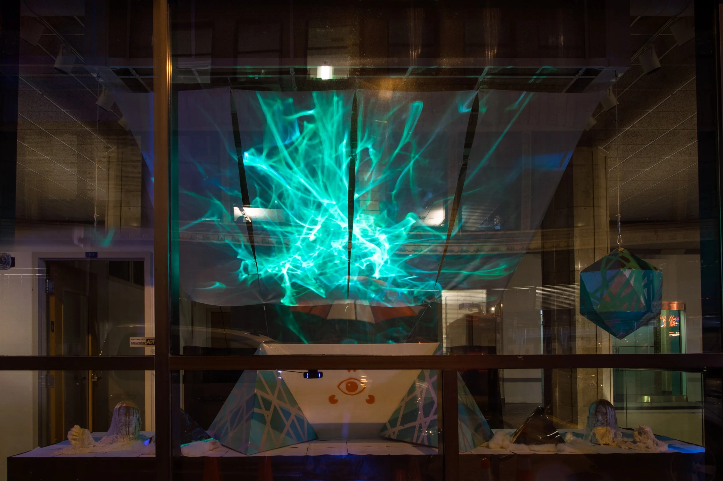 Nighttime street scene reflected on a large window, with a blue and green light display and geometric decorations visible through the window.