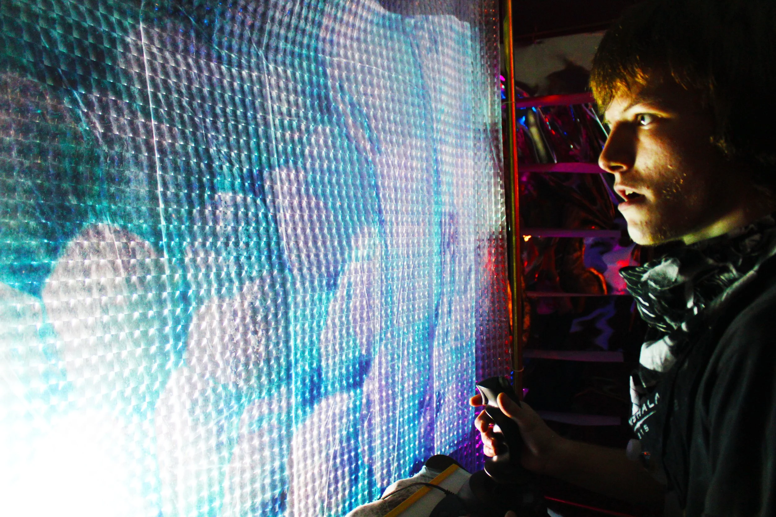 A young man with dark hair and a patterned scarf around his neck playing a digital game on a pixelated, colorful screen in a dimly lit setting.