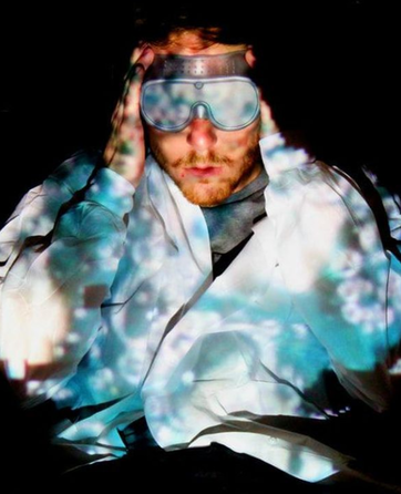 A man wearing ski goggles and a light-colored jacket, with colorful shadows and reflections cast on him, holding the goggles with both hands.