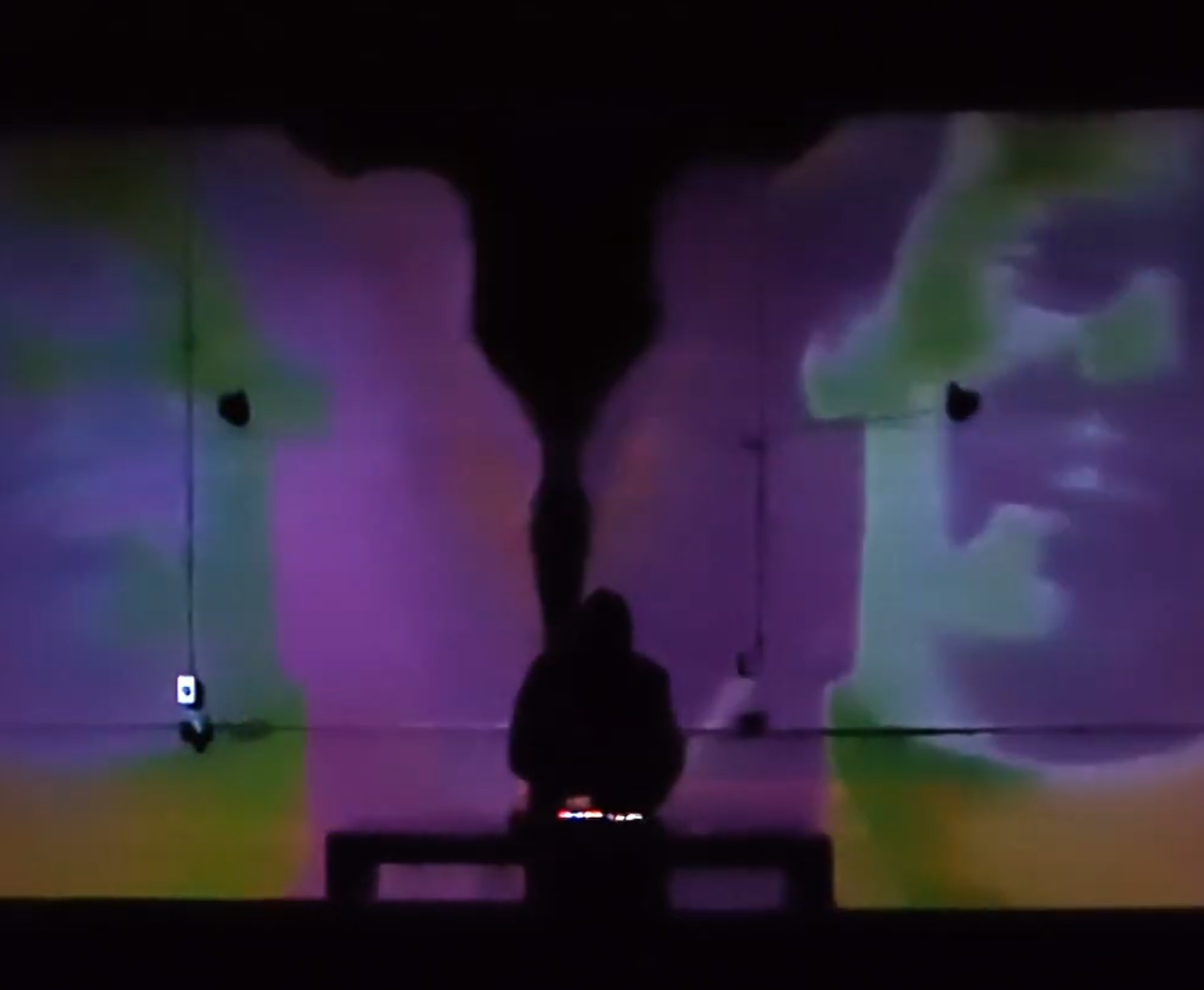 A person with long hair is standing in front of a wall with colorful, abstract light projections. The image is dark, with the person's silhouette visible against the vibrant background.