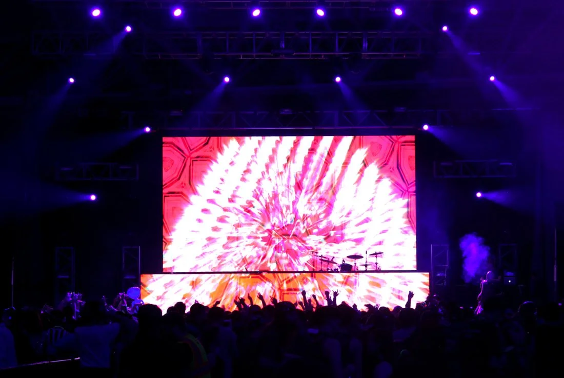 Concert stage with large LED screen displaying bright, colorful visuals and purple stage lighting, with a crowd of people in front.