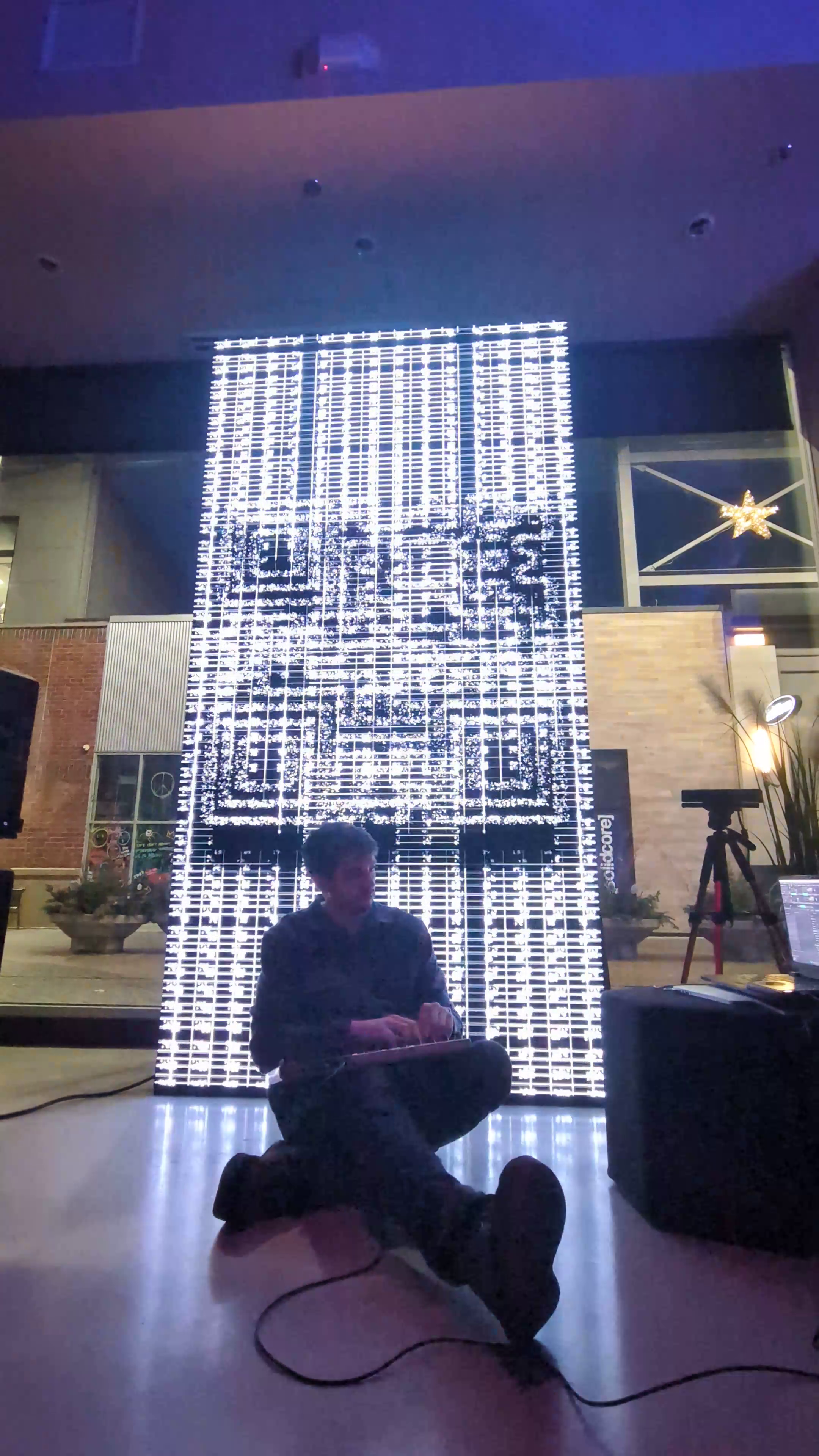 Person sitting cross-legged in front of a large illuminated Christmas display with the message 'Merry Christmas' behind them, indoors.