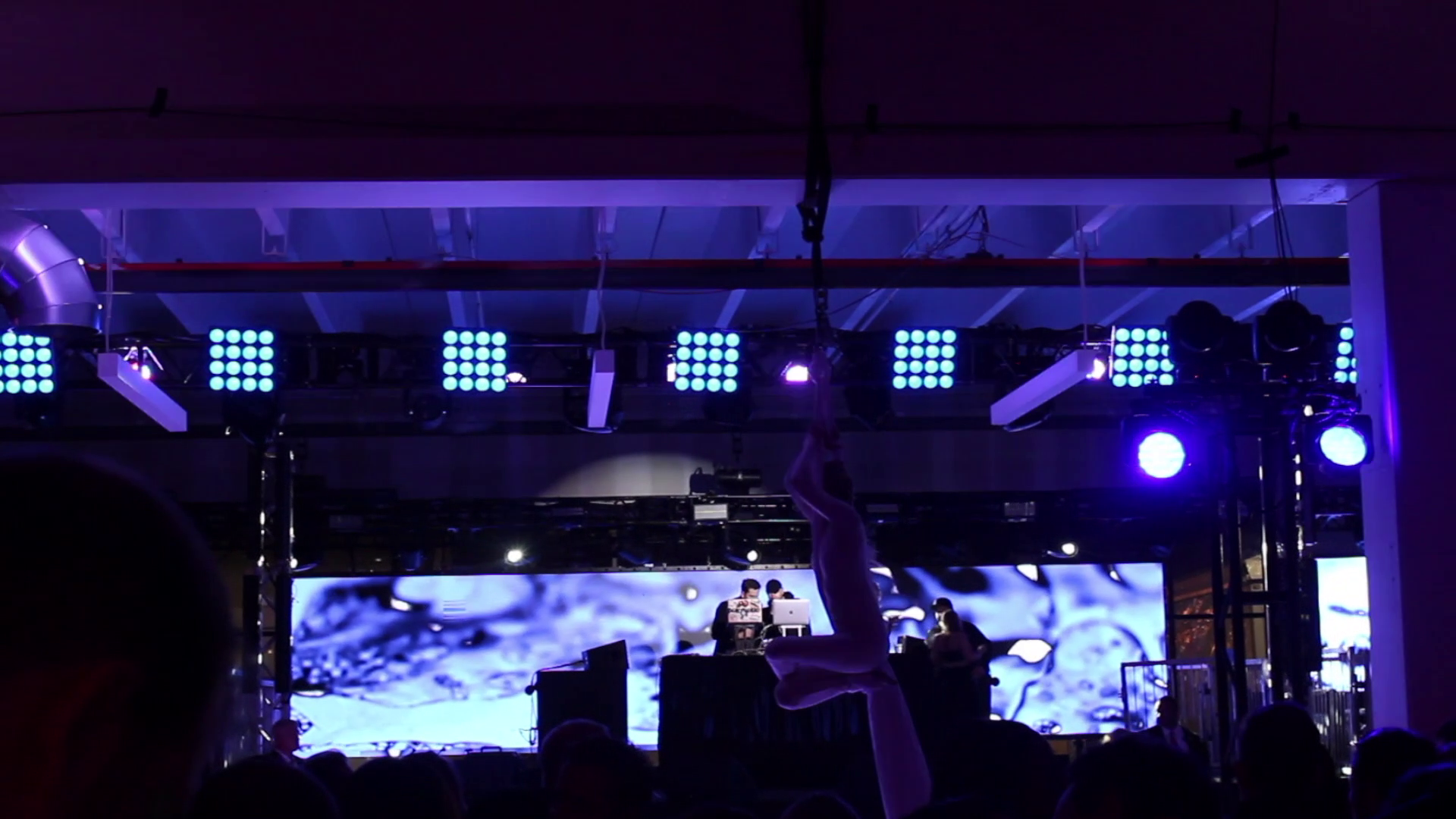 A stage with purple and blue lighting, a large LED screen displaying abstract visuals, and a performer hanging upside down in the center, with an audience in the foreground.