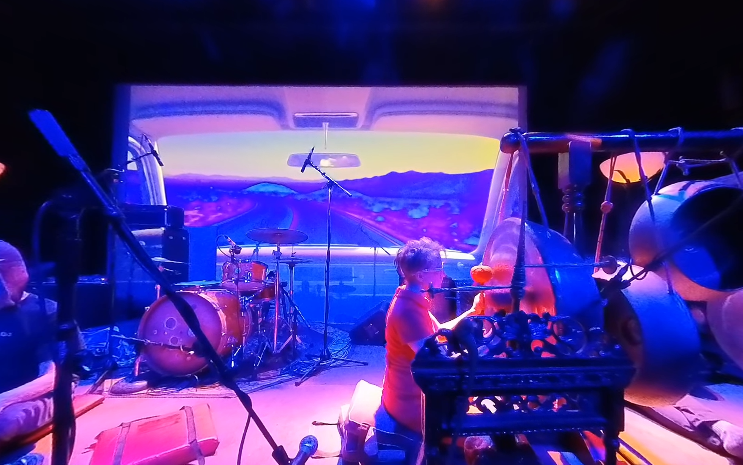 A stage with musical instruments and a large digital screen showing a road scene with mountains at sunset. A person is bending over a cart filled with objects, possibly performing or setting up, with colorful stage lighting.
