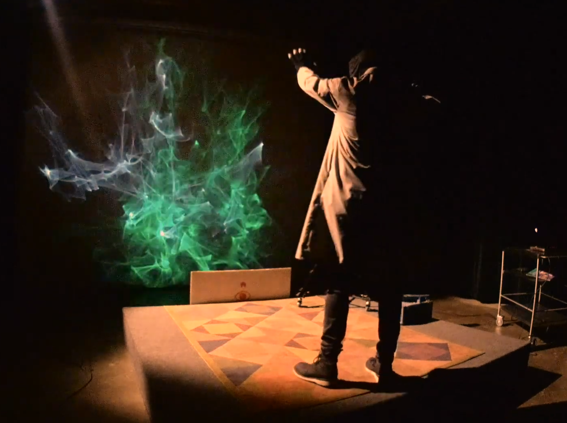 A person on a stage with a patterned rug interacts with colorful smoke. The person is wearing a long coat or dress and is standing on their toes, reaching forward with one arm extended. The smoke appears green and white, swirling in the air against a dark background.