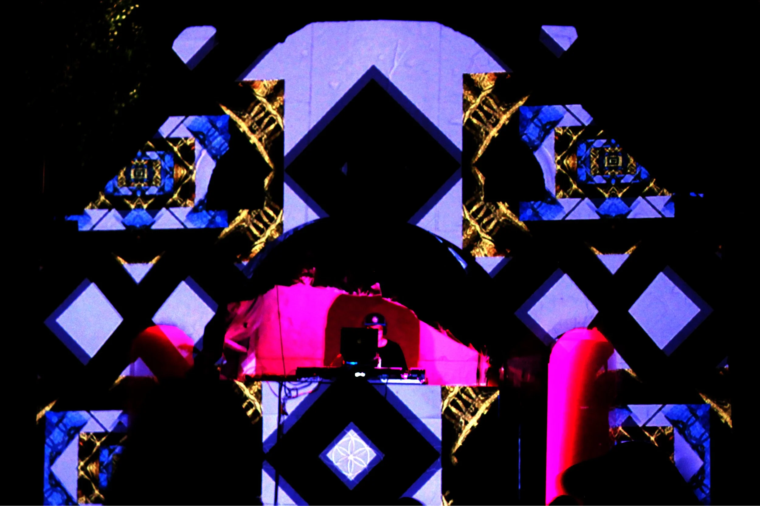A DJ performing at a nighttime event with abstract geometric light projections on a backdrop featuring blue, black, and gold patterns.