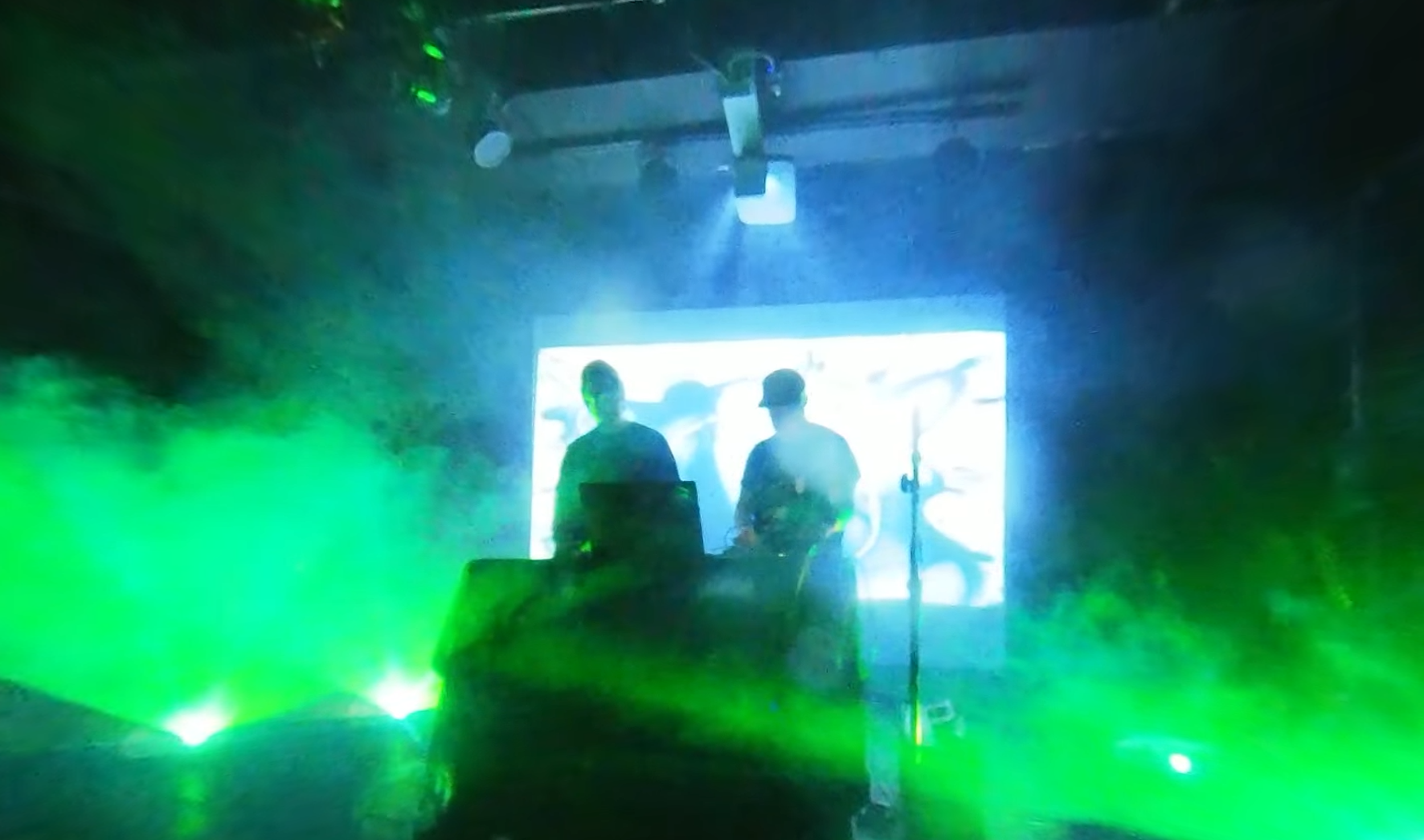 Two DJs perform on a stage with green laser lights and a bright screen in the background, creating a nightclub atmosphere.