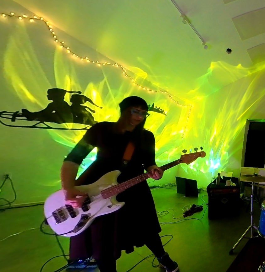 Person playing an electric bass guitar with colorful lights and a silhouette of two children with a dog on a skateboard projected on the wall behind them.