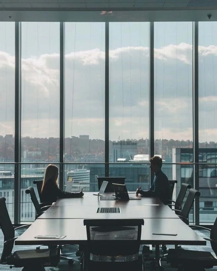 Two people sit at a conference table in a modern office with large windows showing a cityscape and cloudy sky outside.