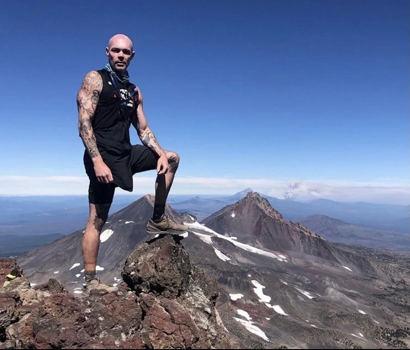 A man with tattoos on his arms, dressed in black athletic gear, standing on a rock with one foot elevated on a smaller rock, on a mountain summit with a volcanic landscape and snow patches, under a clear blue sky.