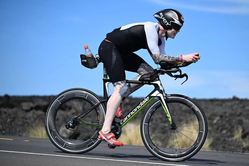 A cyclist wearing a black and white outfit, helmet, and sunglasses riding a Cannondale road bike on a paved road with a rocky landscape and blue sky in the background.