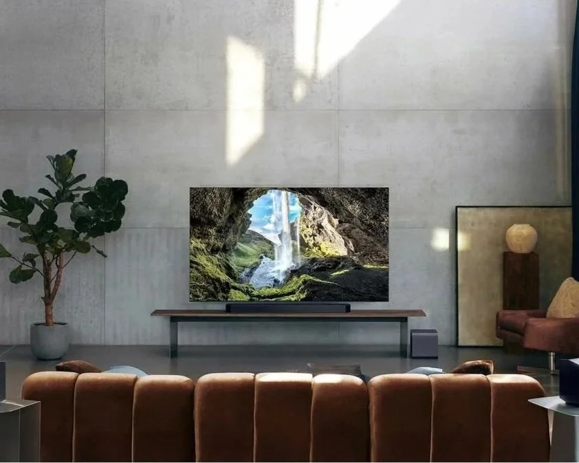 Living room with a large flat-screen TV displaying a waterfall scene, a brown sofa, a potted plant on the left, a pink armchair on the right, a lamp, and a stone wall background with natural sunlight.