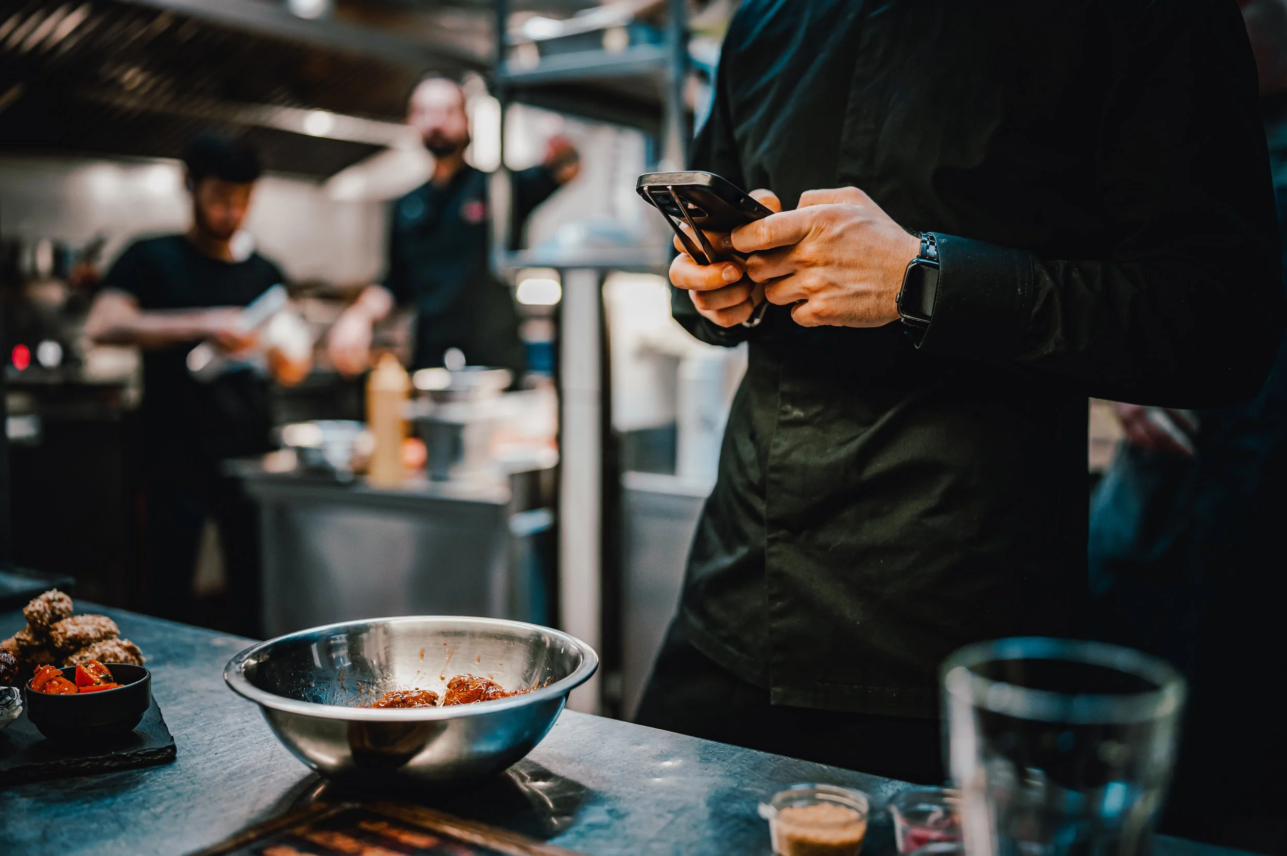 Person in black shirt using a smartphone while cooking in a busy kitchen with two other chefs in the background.