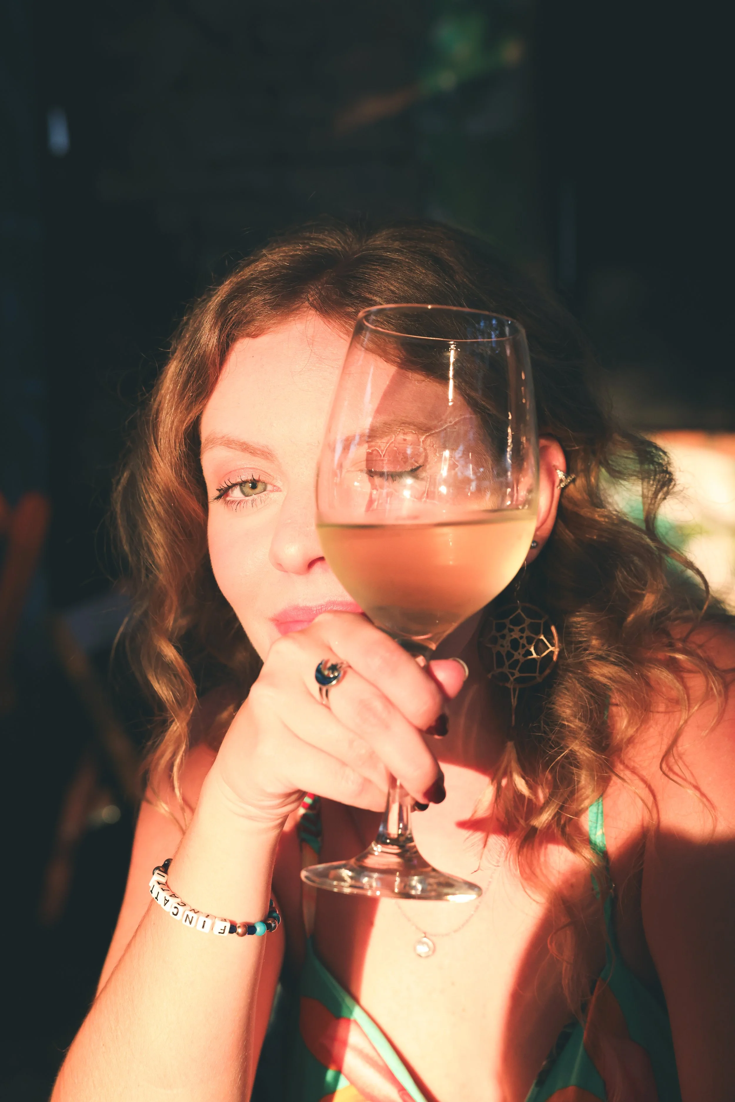A woman with curly hair holding a glass of wine close to her face, winking with one eye, in a dimly lit setting.
