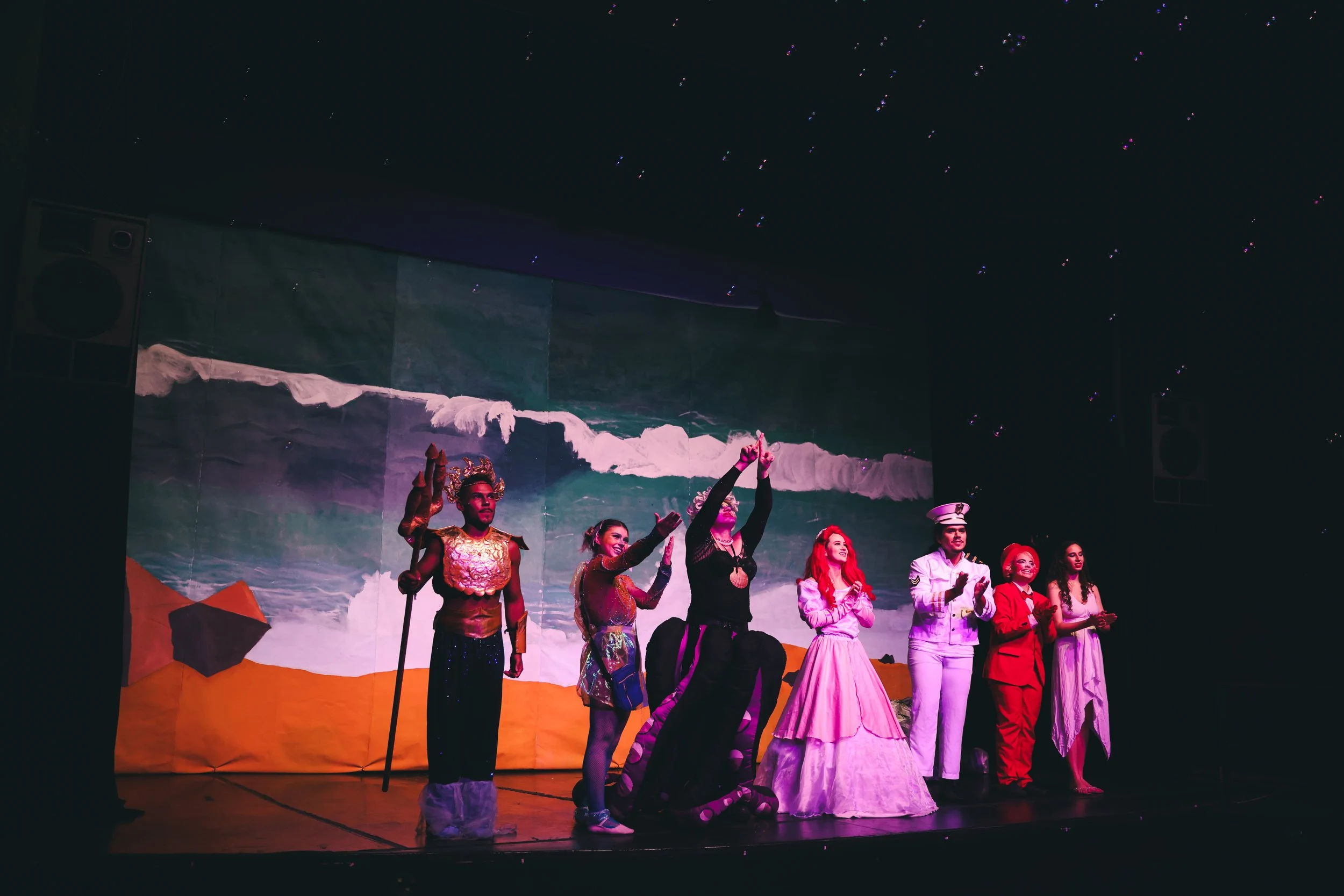 The image shows a stage performance with eight actors in costume, standing in front of a painted backdrop of a beach scene with ocean waves. The actors are dressed in colorful outfits representing different characters, and some are holding their hands up or clapping.