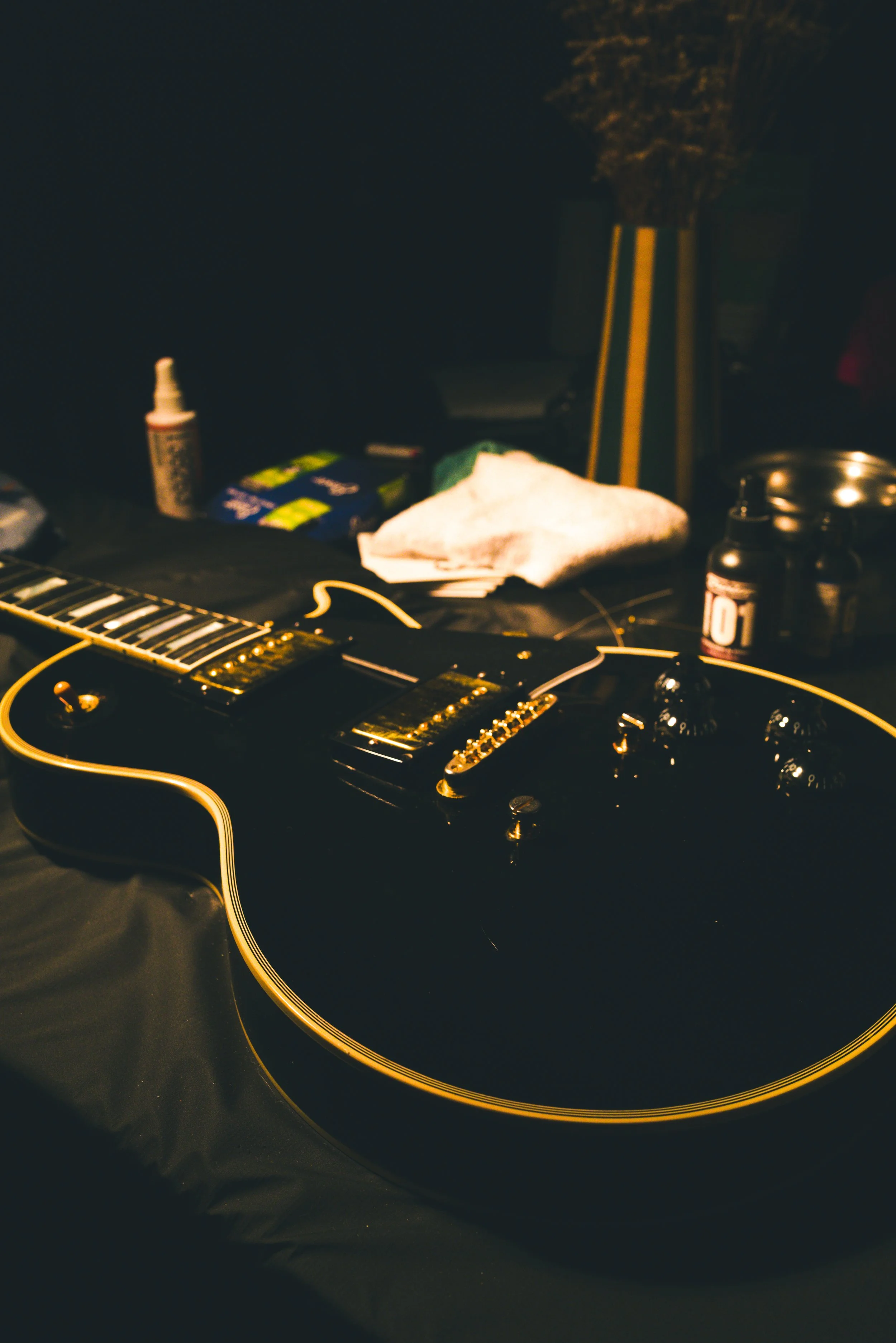 An electric guitar lying on a black surface in a dimly lit room, with guitar effects knobs, and various objects in the background, including a bottle, cloth, and some packaging.